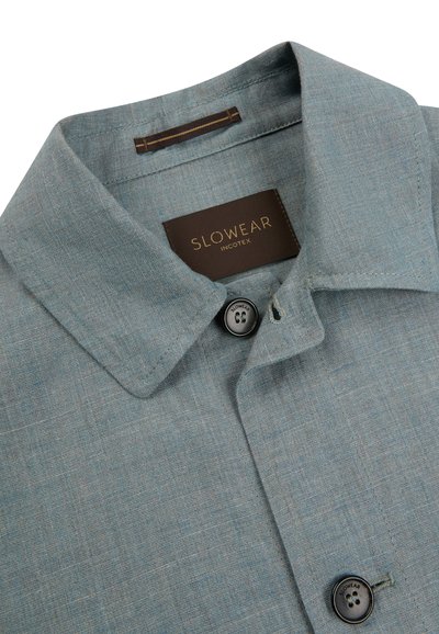 Jasnoniebieski kołnierzyk koszuli z tkaniny o splocie, z czarnymi guzikami z logo "Slowear" oraz brązową metką z napisem "Slowear Incotex" wewnątrz dekoltu.