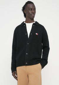 Μαύρη πλεκτή ζακέτα με γιακά σαν ριχτό, με κλείσιμο με κουμπιά και μικρό πολύχρωμο λογότυπο στο αριστερό στήθος. Λεία υφή.