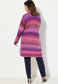 Lange Strickjacke mit lila, pinkem und rotem Farbverlauf; weicher Stoff mit überschnittenen Schultern, getragen über dunklen Hosen und metallischen Knöchelstiefeln.