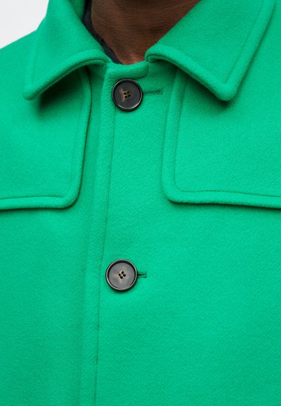 Abrigo de lana verde con cuello, presenta un botón negro y costuras visibles. La tela tiene un aspecto suave y texturizado.