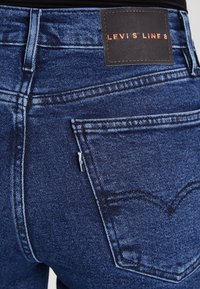 Vaqueros de denim azul oscuro con un diseño distintivo en el bolsillo trasero, parche de logo de cuero y detalles de costura prominentes.