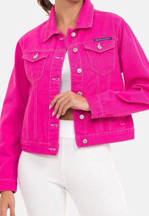Femme portant une veste en denim rose vif avec des boutons argentés sur un crop top blanc côtelé et un pantalon, la main posée près des boutons de la veste.