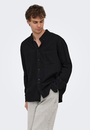Homme portant une chemise noire ample à boutons et un jean gris clair, regardant vers le bas avec une main dans la poche, sur un fond uni.