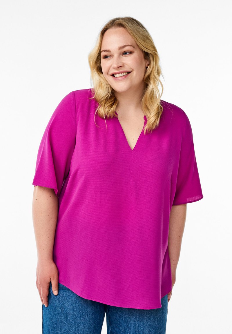 Fuchsia blouse with a V-neck and short sleeves, featuring a relaxed fit. The fabric has a smooth texture, paired with blue denim jeans.