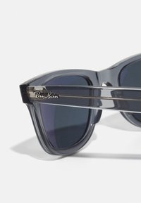 Ochelari de soare gri translucizi cu lentile întunecate, având un logo Ray-Ban pe braț și un design elegant, unghiular.