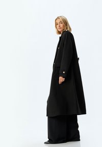 Abrigo negro de gran tamaño con acentos de botones dorados, bolsillos laterales y una textura de lana suave. La modelo lleva unos pantalones anchos negros y zapatos elegantes.