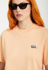 T-shirt en coton p�êche avec un col rond côtelé et un logo noir brodé sur la poitrine. Le modèle porte des lunettes de soleil angulaires noires.