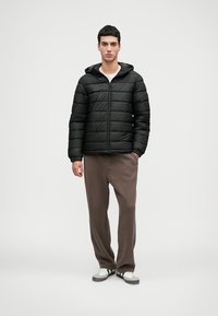 Jachetă puff black cu un model pătrățos orizontal, închidere cu fermoar și glugă. Modelul poartă pantaloni maro și adidași albi, stând în picioare.