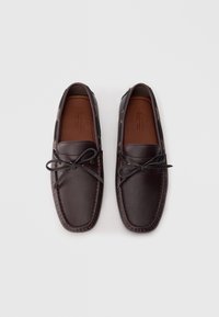 Mocasines de cuero marrón con acabado texturizado, que presentan un lazo atado y ojales dorados, diseñados con una puntera redondeada y suela plana.