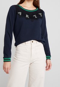 Tmavě modrý top s dlouhým rukávem a zeleným pruhovaným výstřihem, zdobený černou krajkou a nápisem "ARTY" ve flitrové aplikaci na hrudi.