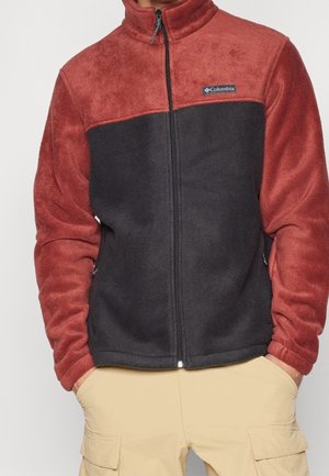 Person trägt eine rot-schwarze Columbia Fleecejacke mit durchgehendem Reißverschluss und beigen Cargohosen, Hände in den Taschen.