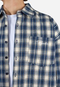 Rutet flannel skjorte i hvid, blå og navy farver; har en knaplukning foran, spids krave og brystlomme med broderet logo.