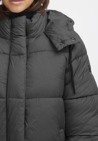 Chaqueta acolchada gris con un patrón de cuadros, cuello alto, capucha y cordón. Cuenta con botones de presión y un tejido texturizado para mayor calidez.