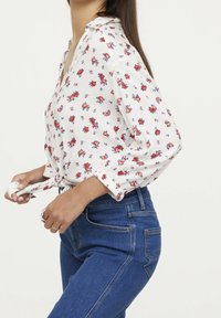 Blusa blanca de flores con flores rojas y azules, fabricada en un tejido ligero, de manga larga y con un nudo en el frente. Combinada con jeans de un azul oscuro.