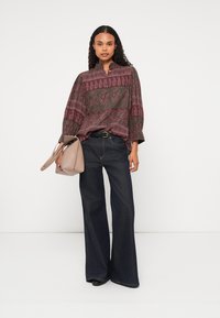 Bordo rakstainā blūze ar augstu apkakli, apvienota ar tumši ziliem platām biksēm. Modelis tur beigu krāsas rokassomu, stāvot pret baltu fonu.