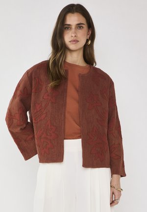 Femme portant une veste brodée de fleurs couleur rouille sur une chemise assortie et une jupe plissée blanche, debout devant un fond uni.
