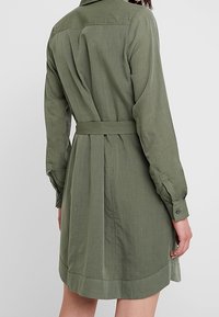 Olivově zelené šaty košilového stylu s dlouhými rukávy, páskem v oblasti pasu a zakřiveným lemem. Vyrobené z lehké látky s viditelnou texturou a detaily šití.