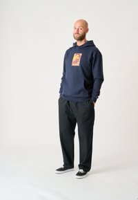 Marineblaues Kapuzensweatshirt mit einer Vordertasche und einem rot-orangenen Grafikdruck mit dem Wort "AHOI". Kombiniert mit schwarzen Hosen und schwarzen Sneakern.