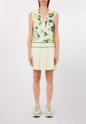 Donna che indossa una maglia senza maniche con colletto e stampa floreale verde e una gonna plissettata color crema abbinata a sneakers beige, in piedi contro uno sfondo bianco.