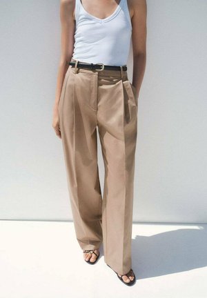 Eine Person trägt ein weißes Trägershirt und beige hochgeschnittene Hosen, kombiniert mit schwarzen Sandalen, und posiert vor einem hellen Hintergrund.