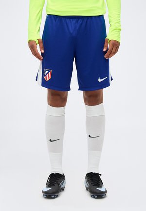 Homme portant un short de football bleu de l'Atlético Madrid, des chaussettes blanches Nike, des crampons noirs et un haut à manches longues vert fluo, debout sur fond blanc.