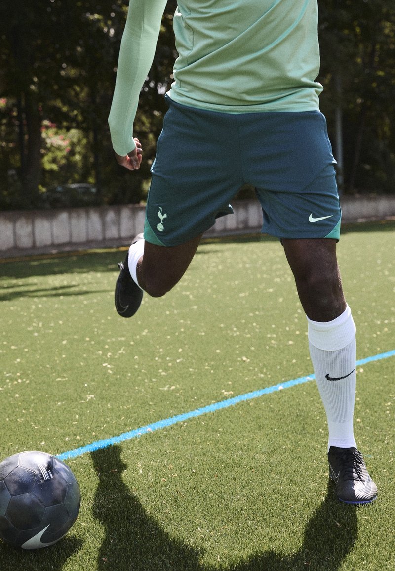 Vihreät Nike-shortsit logolla, mintunvihreä pitkähihainen paita, valkoiset sukat ja mustat jalkapalloilujalkineet. Jalkapallo keinonurmella taustalla.