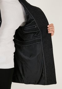 Abrigo de tela exterior texturizada negra con un forro negro suave. El interior cuenta con bolsillos y un detalle de costura en contraste.