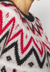 Strickpullover mit weißem Grundton und roten sowie schwarzen Zickzackmustern, runder Ausschnitt und weichem Material. Einzelheiten umfassen kleine schwarze Akzente.