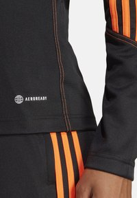 Zwarte sporttop met oranje strepen, voorzien van AEROREADY branding. Gladde textuur en versterkte stiksels langs de zijkanten.