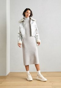 Silvrig metallisk kort jacka med fårskinnskrage, grå ribbad midiklänning, vita strumpor och kraftiga krämfärgade sneakers på trägolv.