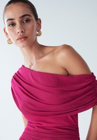 Fuksiansia off-the-shoulder mekko, jossa on varattu kangas ja rypytettyjä yksityiskohtia. Kultaiset teksturoituneet korvakorut täydentävät asua neutraalilla taustalla.