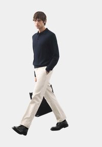 Marineblaue Langarmpolo, cremefarbene Hose, schwarze Anzugschuhe. Trägt eine schwarze Tote-Tasche. Schlichtes, modernes Design mit einfarbigen Tönen.