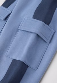 Blauwe stof met verschillende tinten, met een gladde textuur. Twee klepzakken in het bovenste gedeelte voegen detail toe aan het ontwerp.