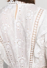 Valkoinen brodeerattu paita, jossa on kukkakuviot, hapsureunus ja napp closure takana avaimenreikää varten.