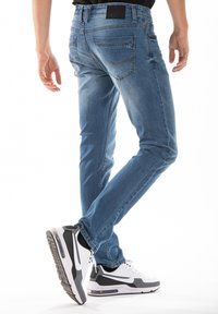 Jean en denim bleu avec une coupe ajustée, comportant des poches arrière et un léger délavage. Associés à des baskets de sport noires et blanches.
