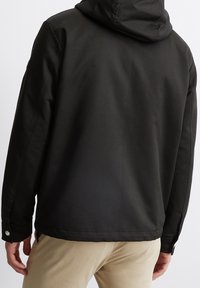 Czarna kurtka z kapturem wykonana z gładkiego, matowego materiału. Posiada boczne kieszenie i mankiety na zatrzaski. Zaprojektowana z luźnym krojem i szwami.