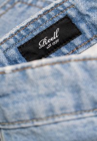 Tejido de mezclilla azul claro con costuras naranjas; presenta una etiqueta negra con texto blanco "Reell est. 1997" en un lugar destacado.