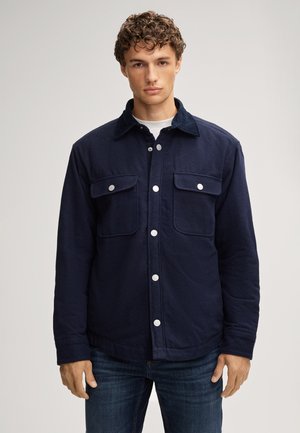 Junger Mann mit lockigem Haar, der eine marineblaue Jacke mit Knöpfen und Klappentaschen sowie blaue Jeans trägt und vor einem einfachen hellen Hintergrund steht.