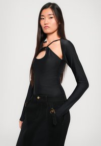 Top preto de mangas longas com um recorte em forma de buraco de fechadura e detalhe de nó no ombro. Textura suave, design ajustado, combinado com uma saia preta de cintura alta.