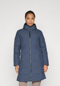 Vrouw in een lange navyblauwe gewatteerde winterjas met capuchon, staande tegen een eenvoudige lichte achtergrond.