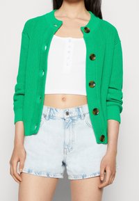 Zelený žebrovaný cardigan s kulatým výstřihem, zapínáním na knoflíky a velkými kontrastními hnědými knoflíky, doplněný bílým topem a světle modrými džínovými šortkami.