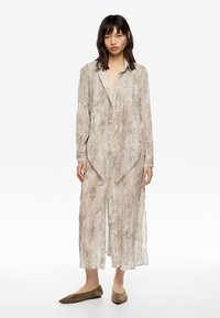 Camisa larga ligera y transparente en color beige con un sutil patrón de serpiente, cierre de botones en la parte delantera y un dobladillo asimétrico; combinada con bailarina suaves de color marrón.