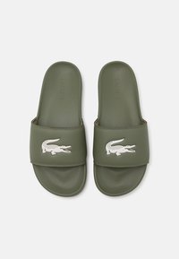 Zöld csúszó szandál sima szintetikus anyagból, fehér Lacoste krokodil logóval a pánton. Lapos kialakítás, sarok nélkül.