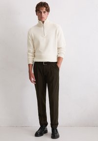 Crème gebreide trui met rits en ribgebreide mouwen, gecombineerd met donkergroene plooibroek en zwarte veterschoenen. Oprichter houding.
