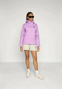 Casaco impermeável roxo claro com capuz e fecho frontal, combinado com calções bege e sapatilhas de cor clara, apresentando um logo no peito.