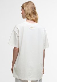 T-shirt de algodão de corte solto e manga curta na cor branca suja, com uma bainha reta e um pequeno detalhe dourado na parte de trás da gola.