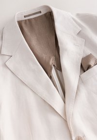 Blazer beige clair en lin texturé, col cranté et doublure en satin à motifs. Comprend une poche poitrine avec un mouchoir assorti.