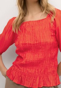 Top rosso a maniche corte realizzato in un tessuto liscio e arricciato, con scollatura quadrata e orlo arricciato, caratterizzato da una texture morbida e una vestibilità rilassata.