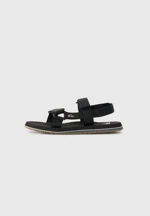 Black sandal featuring adjustable straps made of fabric, a textured flat sole, and a rubber base with a grip pattern.