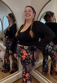 Zwarte wikkel top met lange mouwen en bloemrijke borduursels, gecombineerd met kleurrijke, wijd uitlopende broek met diverse patronen en texturen.